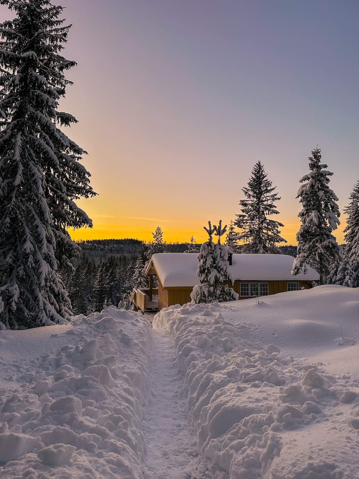 A winter scene showcases a cozy cabin nestled among snow-covered trees, with a pathway leading to the entrance. The horizon is painted with soft hues of yellow and orange from the setting sun, creating a serene atmosphere in the tranquil landscape.