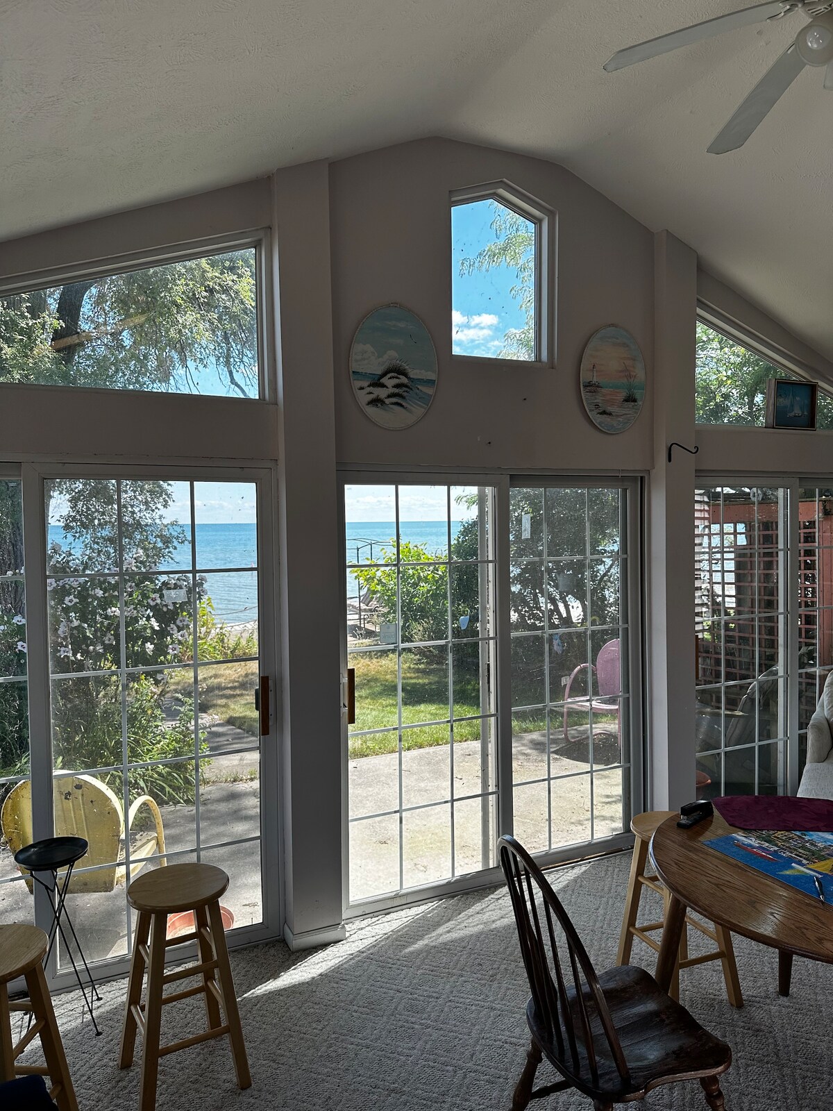 Large glass doors allow natural light to fill the space, providing a view of Lake Huron. The interior features wooden furniture, including a round dining table. Colorful wall art and a mix of seating options create a welcoming environment.