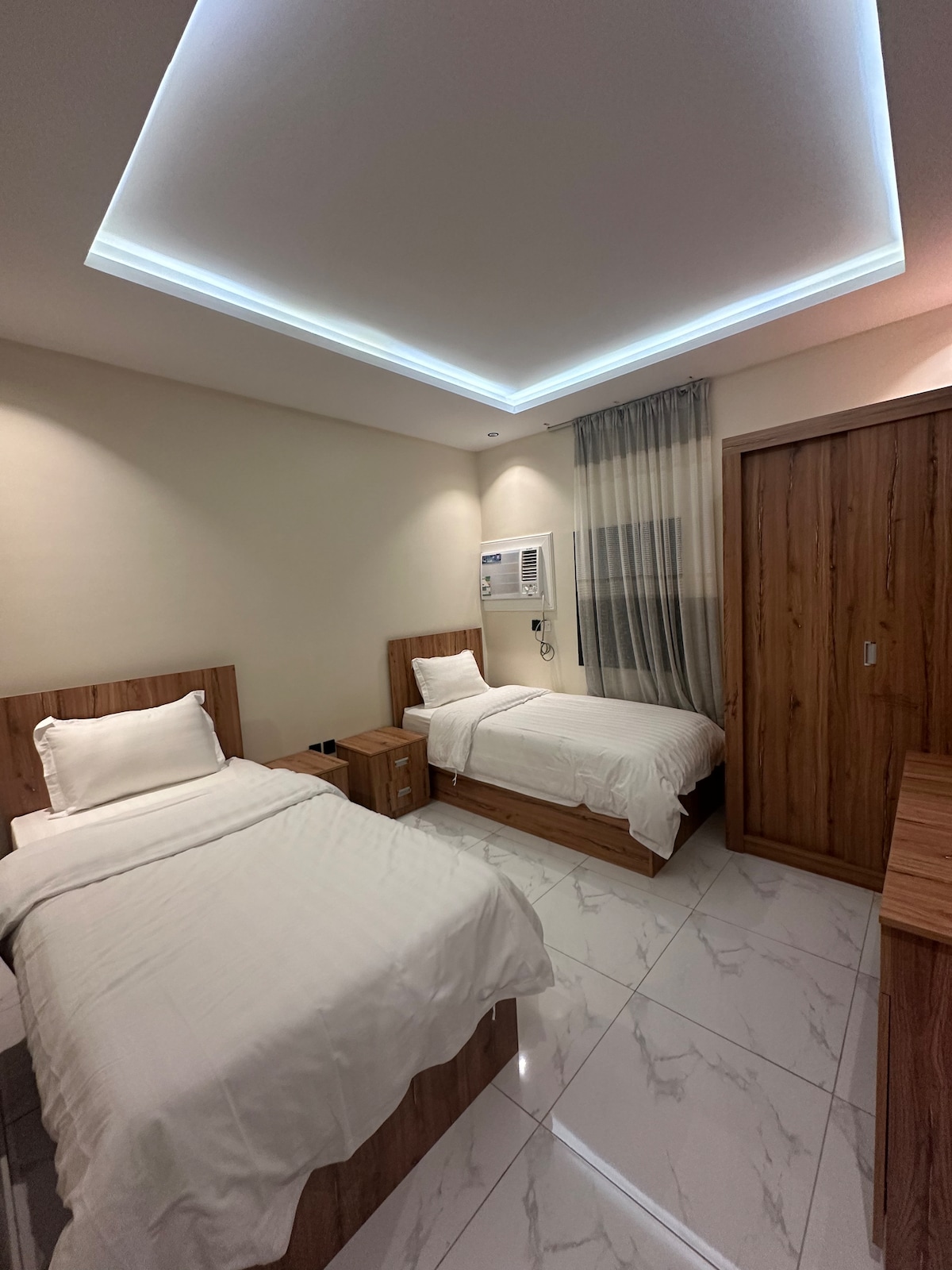 The room features two separate beds with light-colored linens, positioned against the wall opposite a wooden wardrobe. A minimalistic design is complemented by a soft, illuminated ceiling, and a compact air conditioning unit is mounted for comfort. Natural light is softly filtered through sheer curtains.