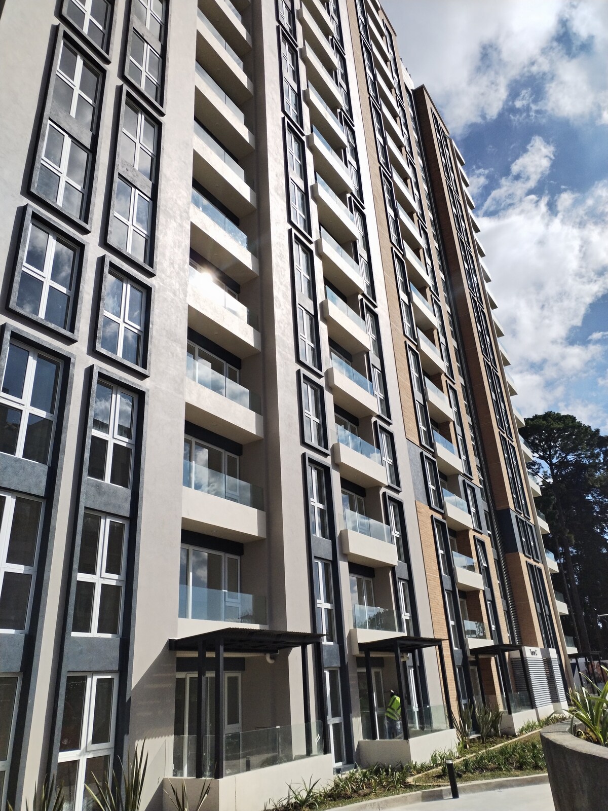 The exterior of a modern multi-story building is displayed, featuring a blend of light and dark facade panels. Multiple balconies are visible, with glass railings enhancing the contemporary design. The surroundings include greenery, offering a connection to nature.