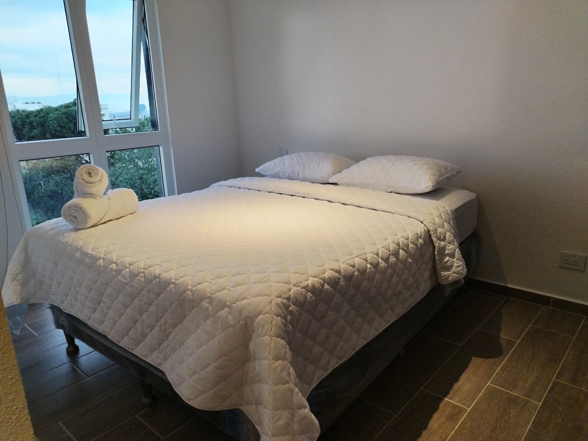 A spacious bed with a white quilted cover and two neatly arranged towels is positioned against a large window. Light enters the room, creating a bright and airy atmosphere. Greenery is visible outside, enhancing the serene ambiance.