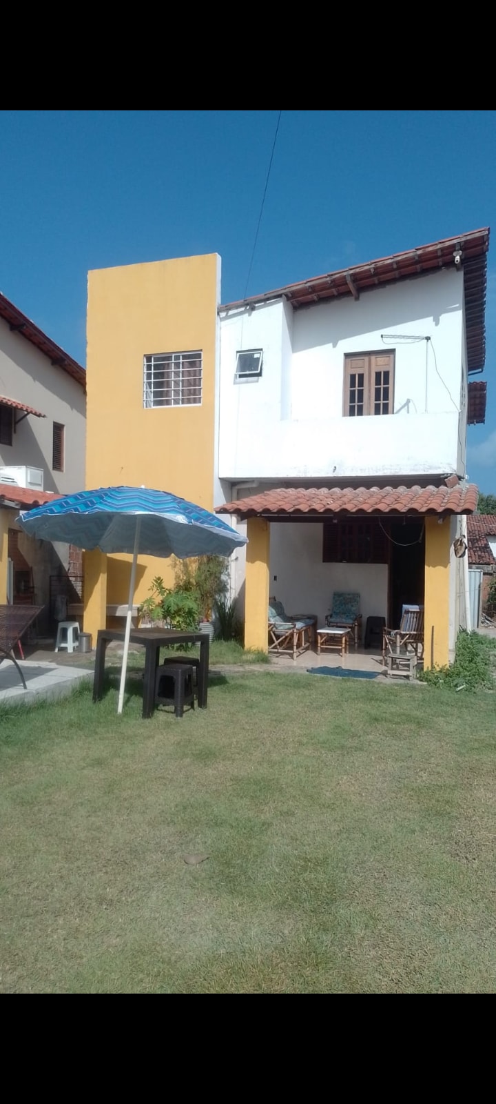 The exterior of a two-story house is visible, featuring a yellow wall and a tiled roof. A shaded area with outdoor seating is present, alongside a table and chairs on the lawn. Clear blue sky enhances the inviting atmosphere of the spacious yard.