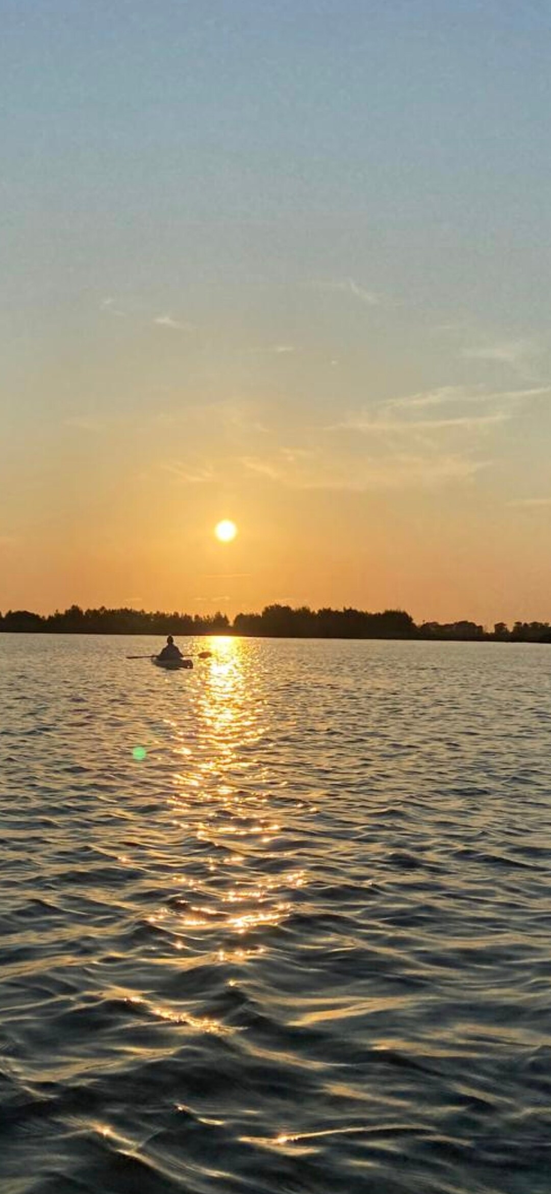 A serene sunset illuminates the sky with warm hues, reflecting on the water's surface. A person in a kayak is visible, gliding gently across the lake, surrounded by silhouettes of trees in the background.