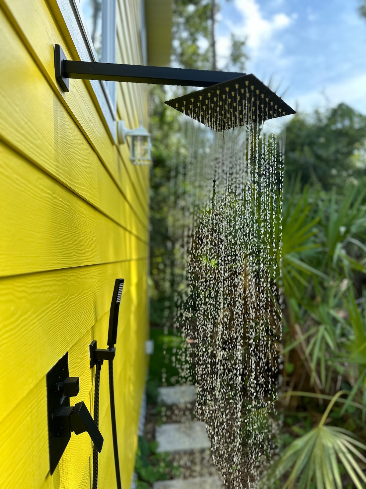 An outdoor shower is mounted on a vibrant yellow exterior wall, featuring a square showerhead from which water droplets cascade. Lush greenery is visible in the background, complemented by a stone pathway leading away from the shower area.