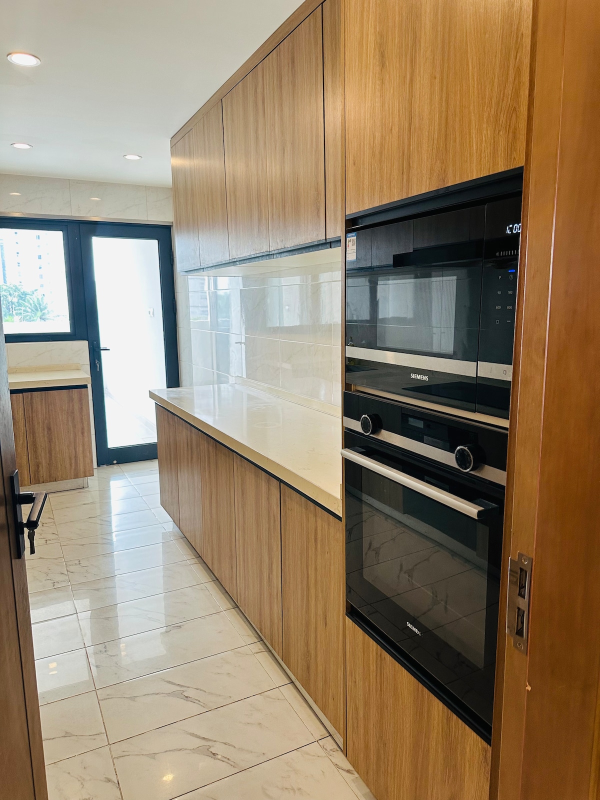 The kitchen features sleek wooden cabinetry, equipped with modern appliances, including an oven and microwave. Light-colored countertops complement the design, while large windows allow natural light to fill the space. A minimalist aesthetic is maintained with a clean and organized layout throughout.