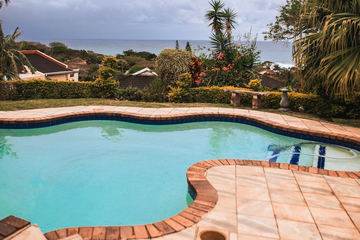The outdoor pool features a curved shape and is surrounded by tiled decking. Lush greenery and colorful flowers frame the area, while glimpses of the ocean are visible in the background under a cloudy sky.