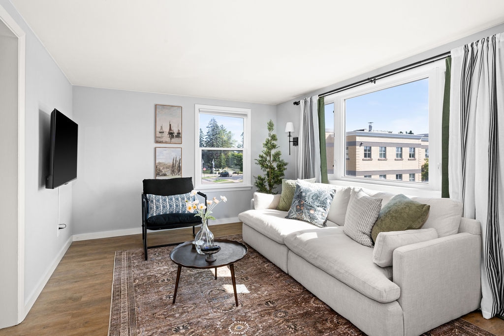 A bright living area features a spacious, light-colored sofa complemented by decorative pillows. A small round table sits in front, adorned with a vase containing fresh flowers. Natural light enters through two windows, framed by green curtains, and artworks decorate the walls.