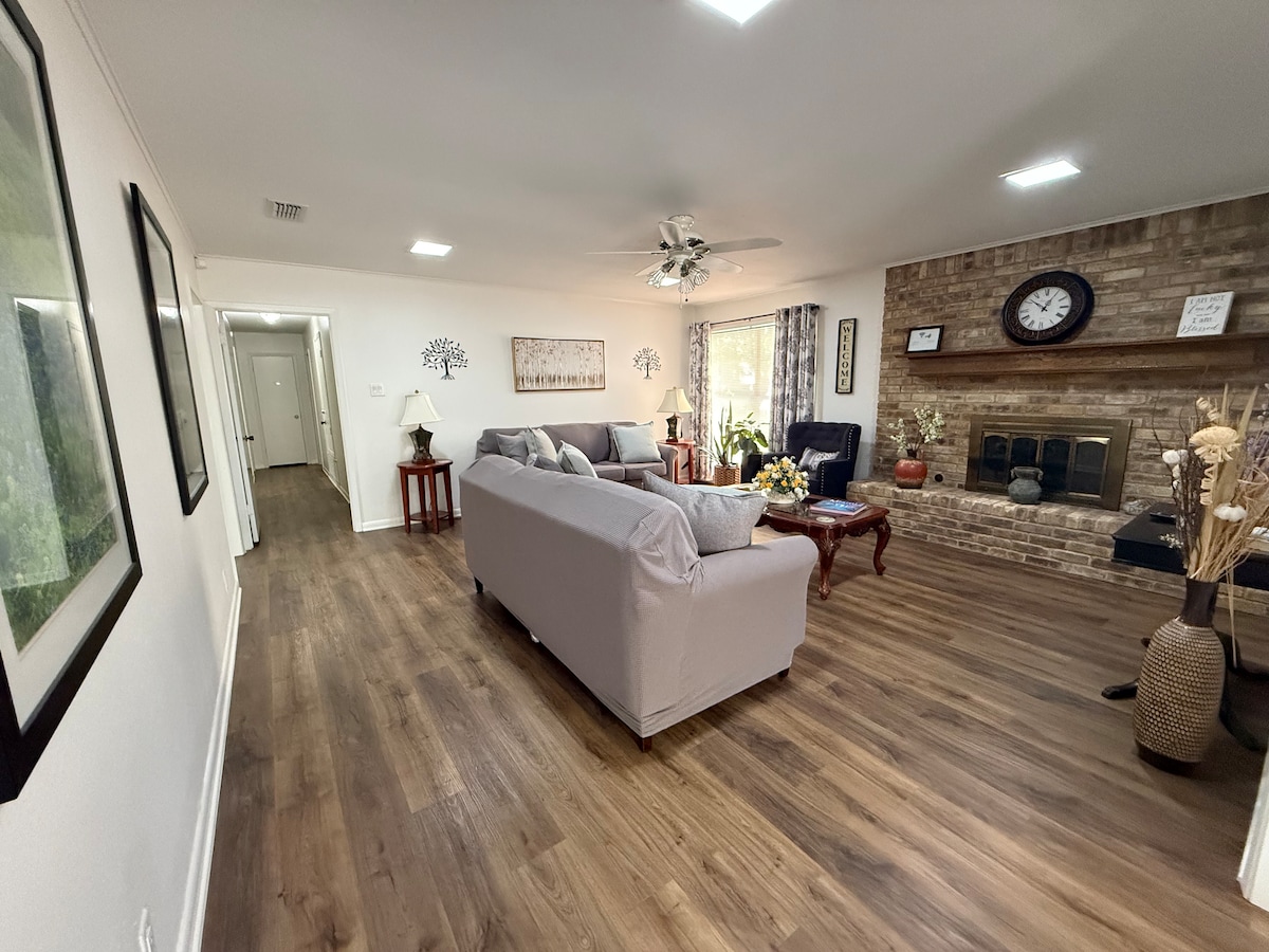 A welcoming living area features a comfortable light-colored sofa facing a brick fireplace. Light wooden flooring extends throughout the space. Large windows allow natural light to fill the room, while decorative elements and wall art enhance the cozy ambiance.