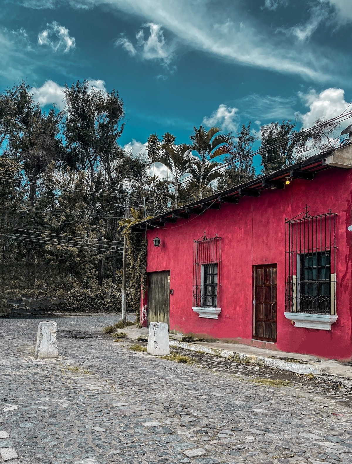 A vibrant red building is framed by lush greenery under a blue sky with scattered clouds. Cobblestone streets intersect nearby, adding to the charm of the colonial setting. The building features black wrought iron bars on windows and a wooden door, creating a classic aesthetic.