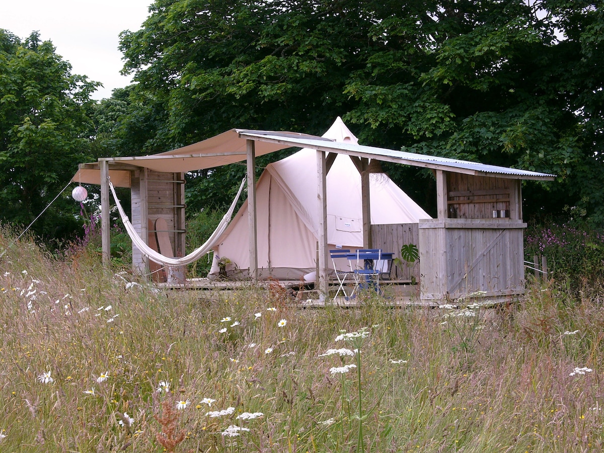 A bell tent is situated on a wooden platform, surrounded by wildflower meadows. A hammock is suspended from the structure, while a small table and chair set is positioned nearby. Natural greenery and trees frame the peaceful outdoor setting.