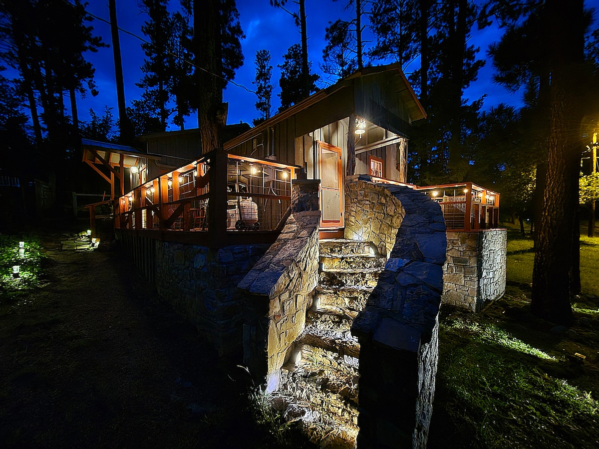 A charming stone staircase meanders up to a warmly lit cabin nestled among tall trees, creating an inviting appearance during twilight. Outdoor seating areas with wooden railings are visible, enhancing the rustic ambiance of the space.