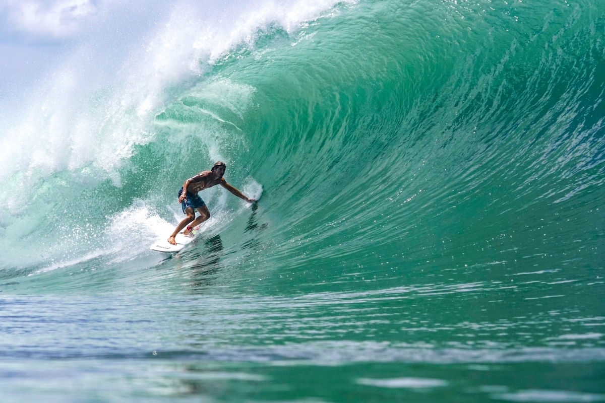 A surfer is seen riding a large green wave, showcasing dynamic motion. The water's surface reflects varying shades of turquoise, contrasting with the white crest of the wave. In the background, a clear sky enhances the vibrant colors of the scene.