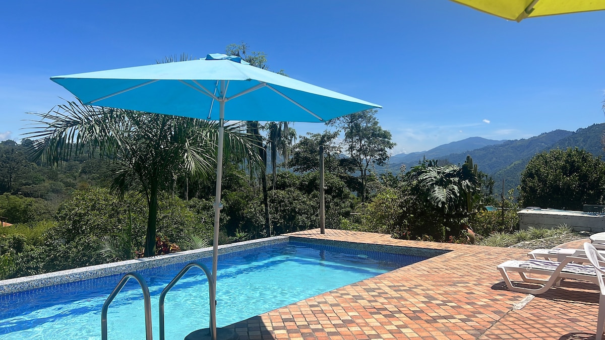 A clear blue pool invites relaxation, surrounded by lush greenery and rolling hills. Two umbrellas provide shade, while white lounge chairs are arranged nearby. The distant mountains create a serene backdrop under a bright blue sky.
