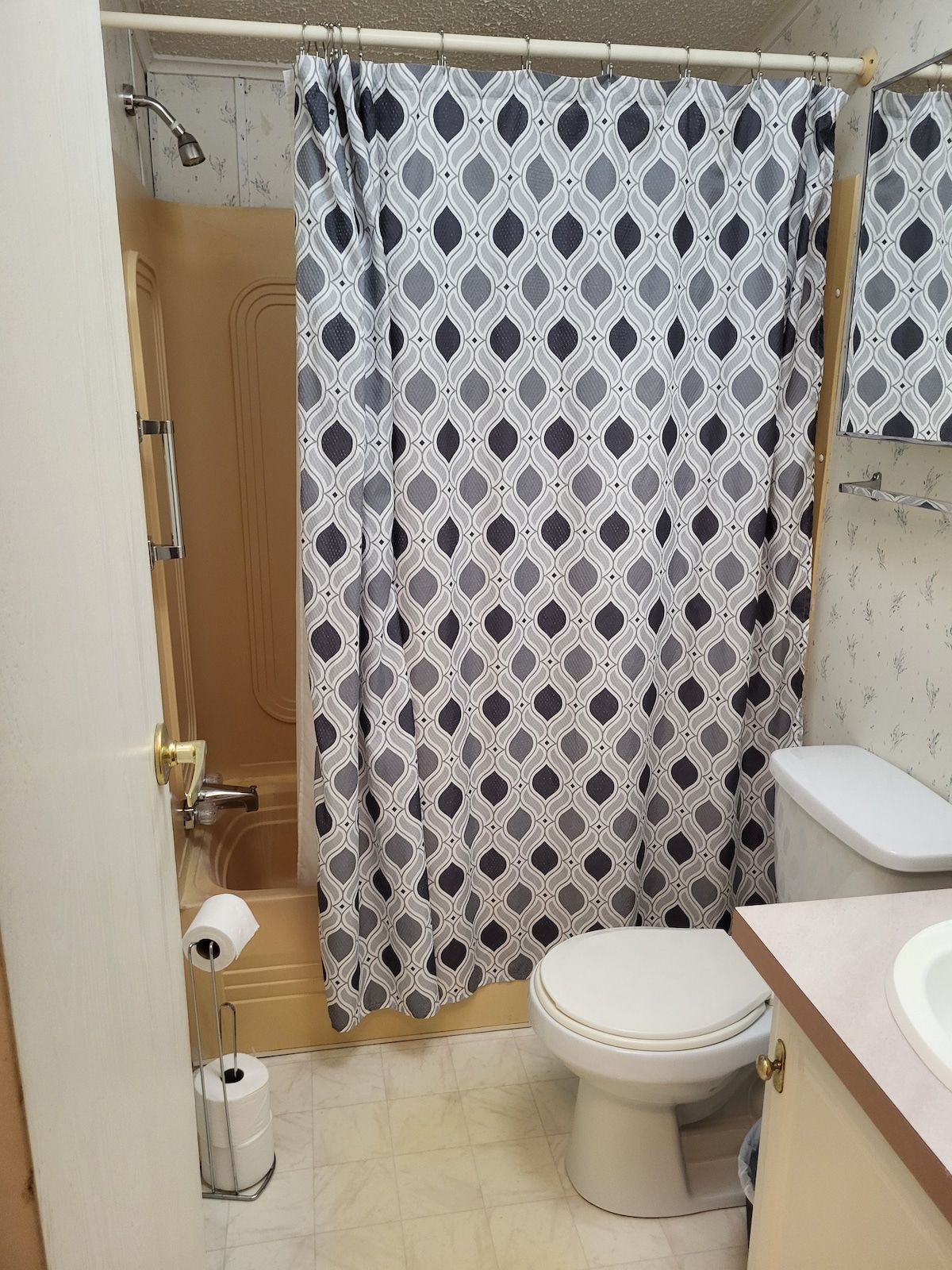 A compact bathroom is shown, featuring a neutral colour scheme. A walk-in shower with a patterned curtain is visible alongside a toilet and a small sink with a light countertop. Essentials like toilet paper and hand towels are neatly arranged for convenience.