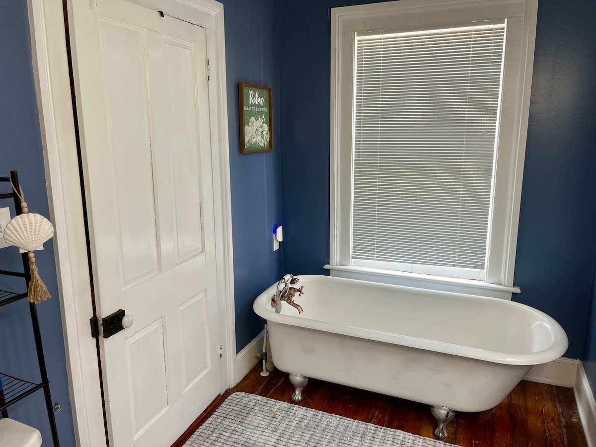 The en suite bathroom features a classic clawfoot bathtub against a blue wall. A white window with blinds allows natural light to fill the space. A textured rug is positioned on the wooden floor, and a decorative wall piece complement the simple decor.