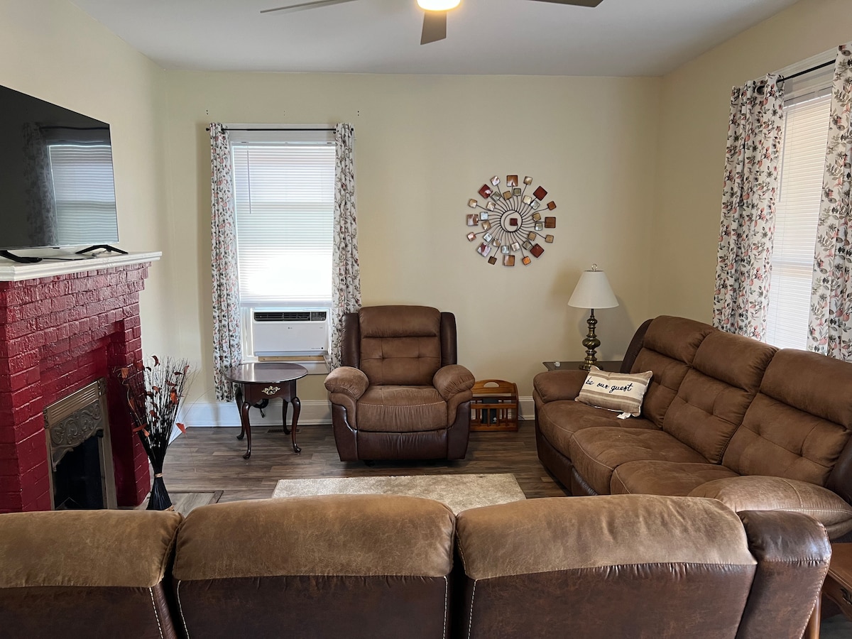 A comfortable living area is displayed, featuring a large sectional sofa and two reclining chairs. A TV is mounted above a decorative brick fireplace. Natural light enters through two windows covered with floral curtains, while a small side table and a lamp complete the space.