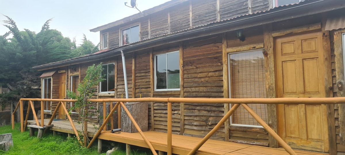A rustic wooden cabin is surrounded by lush greenery, showcasing a wraparound wooden deck with sturdy railings. Large windows offer views into the cozy interior, while the front door features a simple design, adding to the inviting exterior of the accommodation.