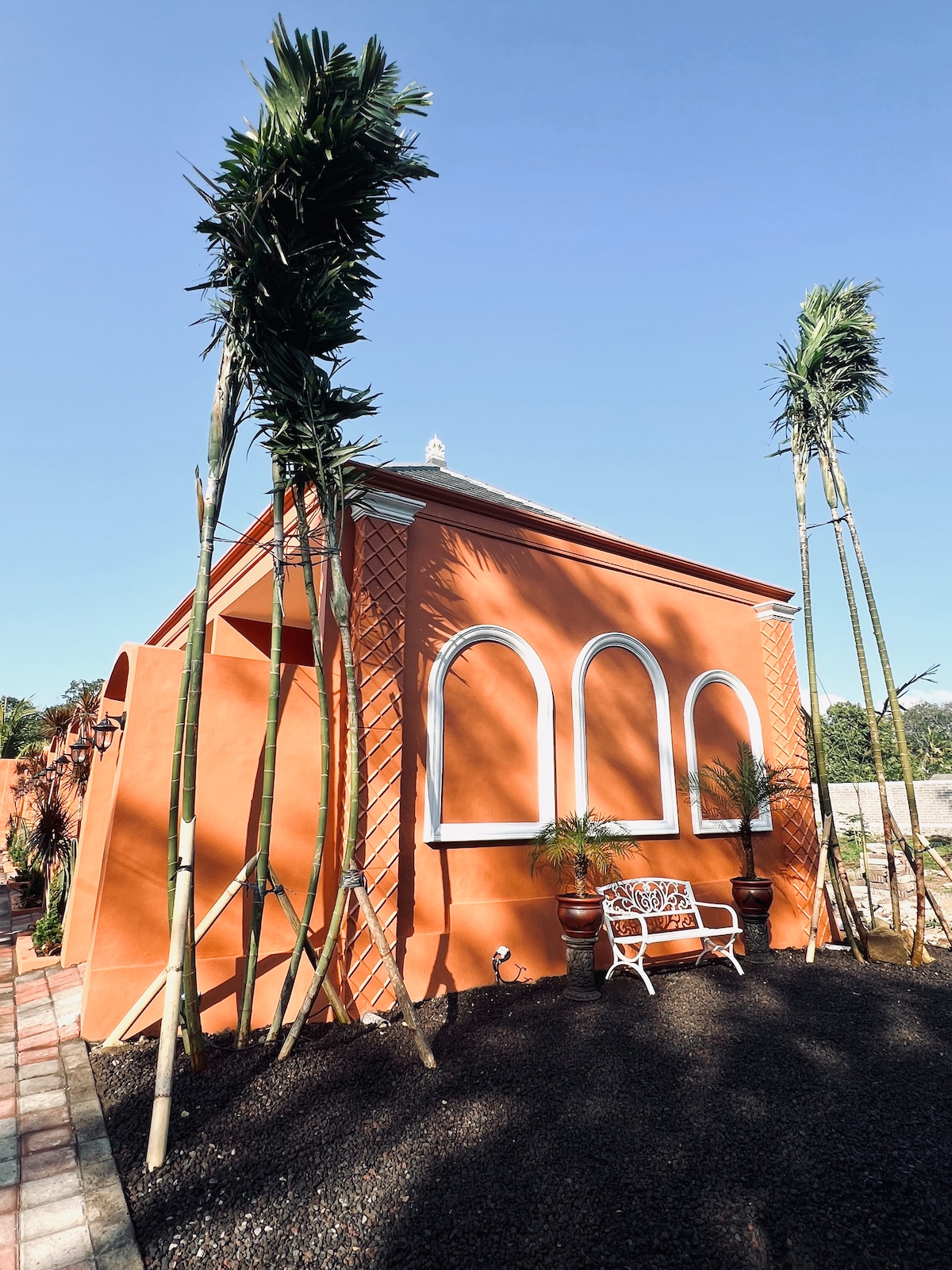 A vibrant orange exterior is showcased, featuring large arched windows framed in white. Lush palm trees enhance the tropical feel, while a small white bench accentuates the inviting entryway. The surrounding area is landscaped with dark gravel, creating a neat and welcoming atmosphere.
