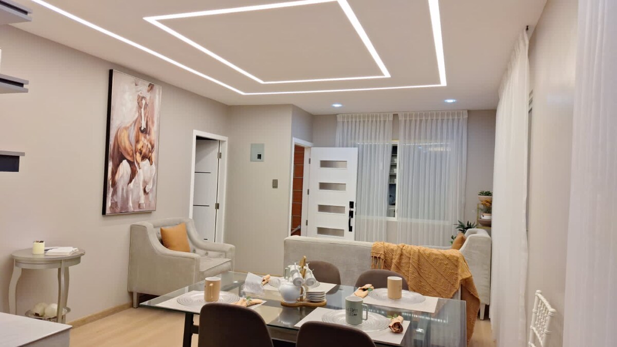 A modern living space is displayed with soft neutral tones and contemporary furniture. A decorative painting of horses adorns the wall. Natural light filters through sheer curtains, illuminating a dining area with a stylish table set for breakfast, featuring plates and cups.