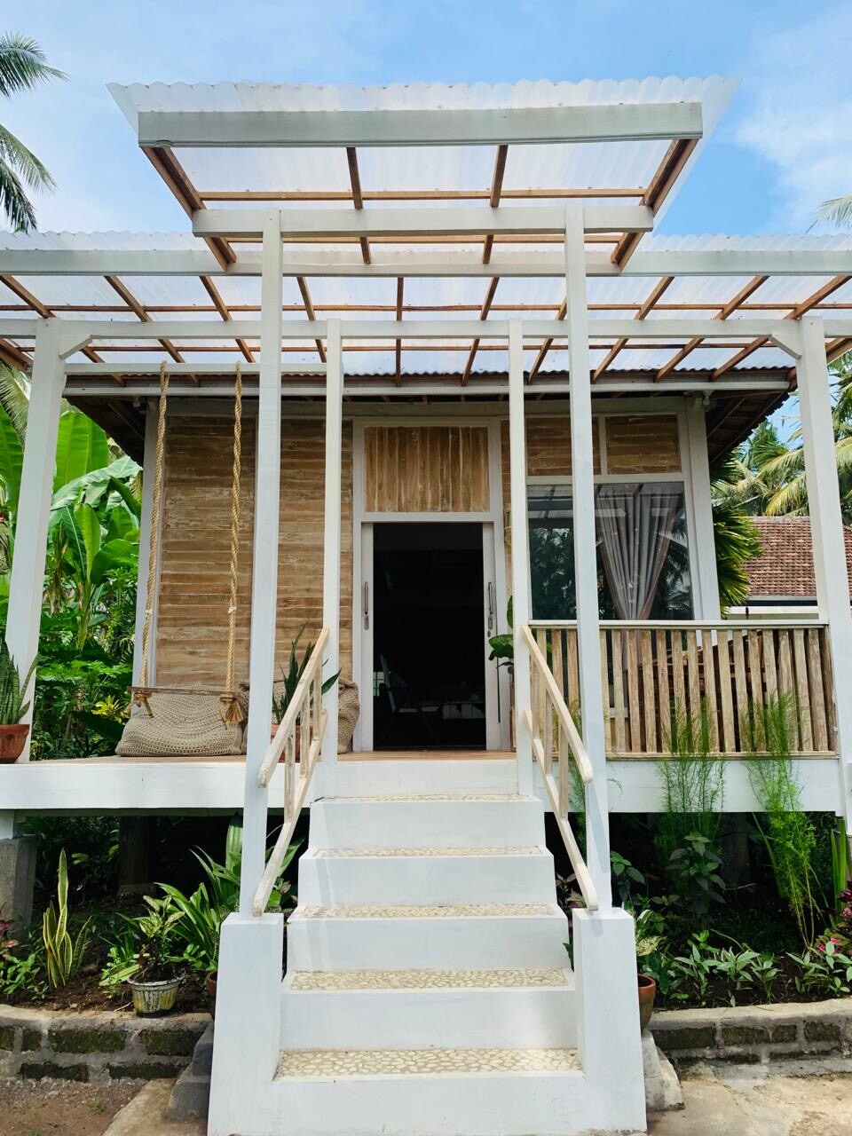 A charming wooden home is showcased with a welcoming entrance featuring white steps leading up to a covered porch. The exterior is surrounded by lush greenery, with a swing hanging from the porch and potted plants enhancing the natural setting.
