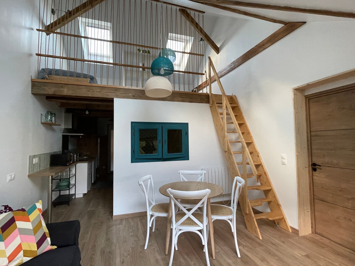 The living area features a light wooden staircase leading to a mezzanine level. A round table with four white chairs sits in the foreground, surrounded by neutral-toned decor. Natural light filters through the skylights, enhancing the spacious feeling of the room.