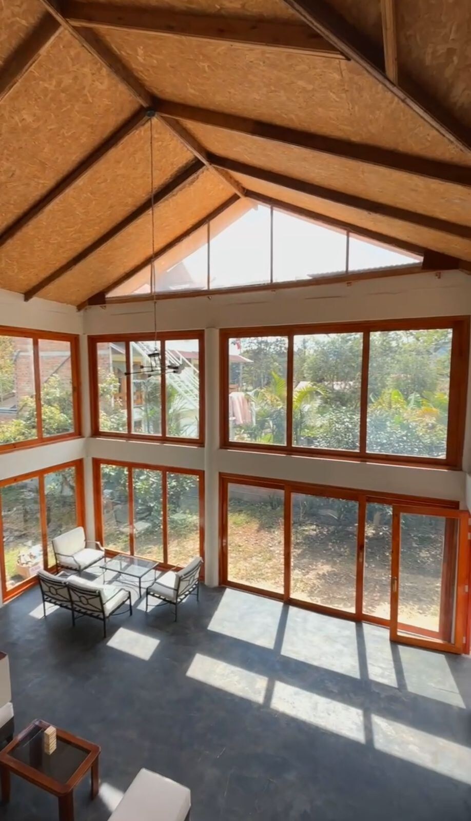 A sunlit common area is defined by tall windows framing views of the surrounding foliage. Natural light streams in through the glass, creating a warm ambiance. A seating arrangement of two chairs and a table is situated in the center, inviting relaxation.