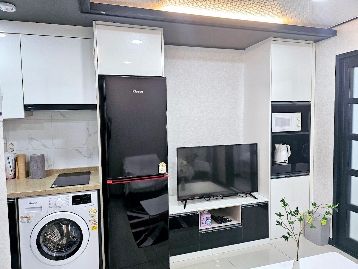 A modern kitchen area features sleek white cabinets and black appliances. A washing machine is visible alongside a refrigerator and microwave. A wall-mounted TV is positioned above a low cabinet, while a small plant adds a touch of greenery to the space.