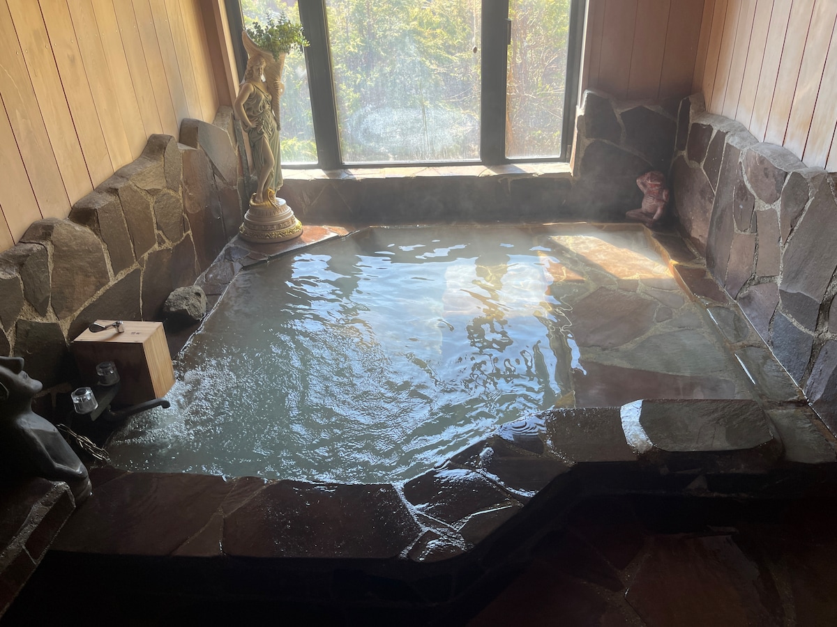 A tranquil hot spring bath is framed by natural stone walls and features a wood and stone bench. Sunlight filters through a large window, casting reflections on the water's surface. Decorative elements include a small statue and a wooden box beside the bath.
