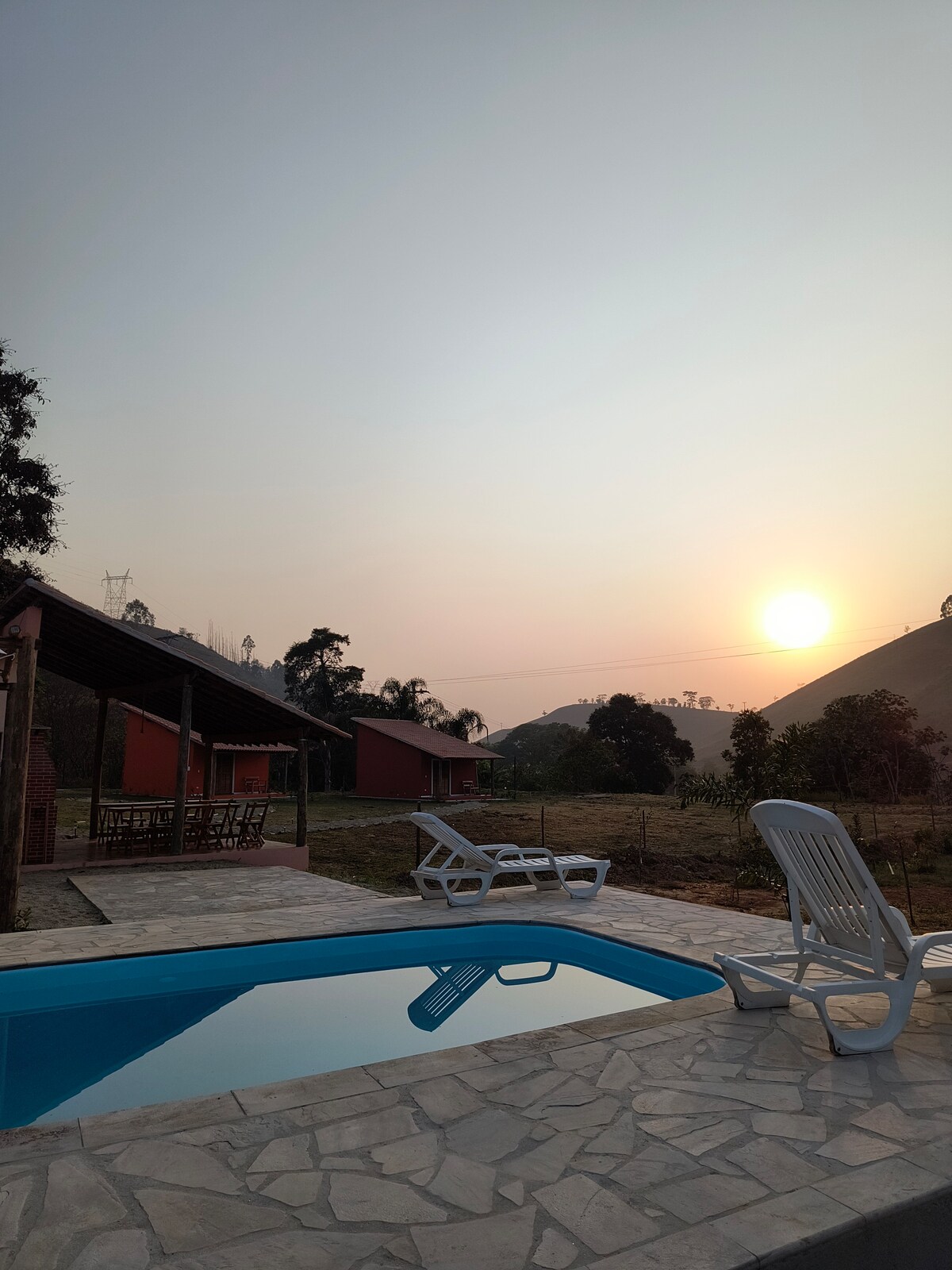 A tranquil scene features a swimming pool reflecting the early morning light, surrounded by paved stone and lounge chairs. In the background, rustic chalets are positioned against rolling hills, while the sun rises gently beyond the landscape.