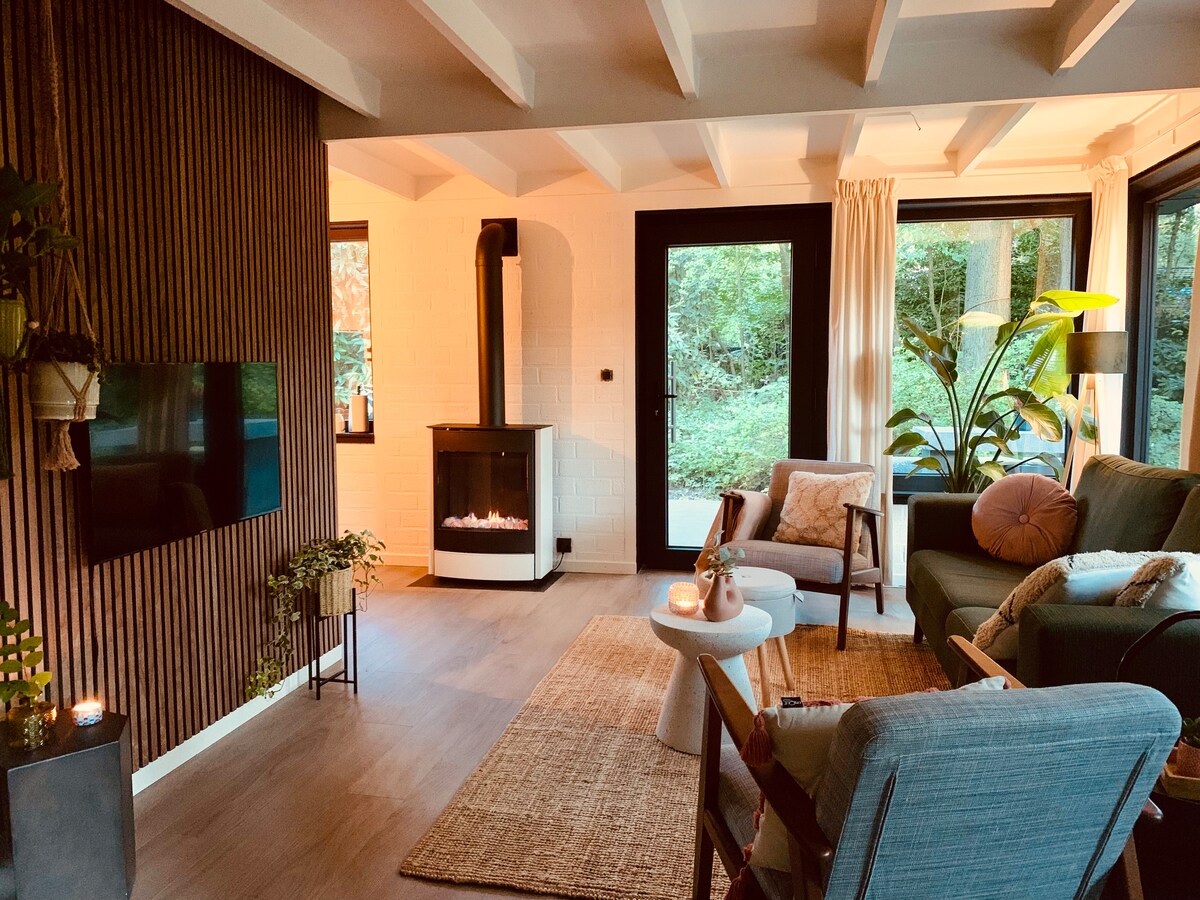 A cozy living area features a stylish propane fireplace against a wooden accent wall. Large windows frame views of the surrounding greenery. A comfortable seating arrangement includes a green sofa and a light blue armchair, complemented by a circular coffee table and a woven rug.