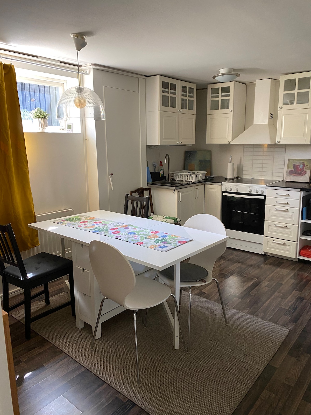 A well-equipped kitchen features white cabinetry and a stove. A small dining table is surrounded by white and black chairs. Natural light filters through a nearby window, with a yellow curtain adding a pop of color. A neutral rug complements the dark wooden floor.