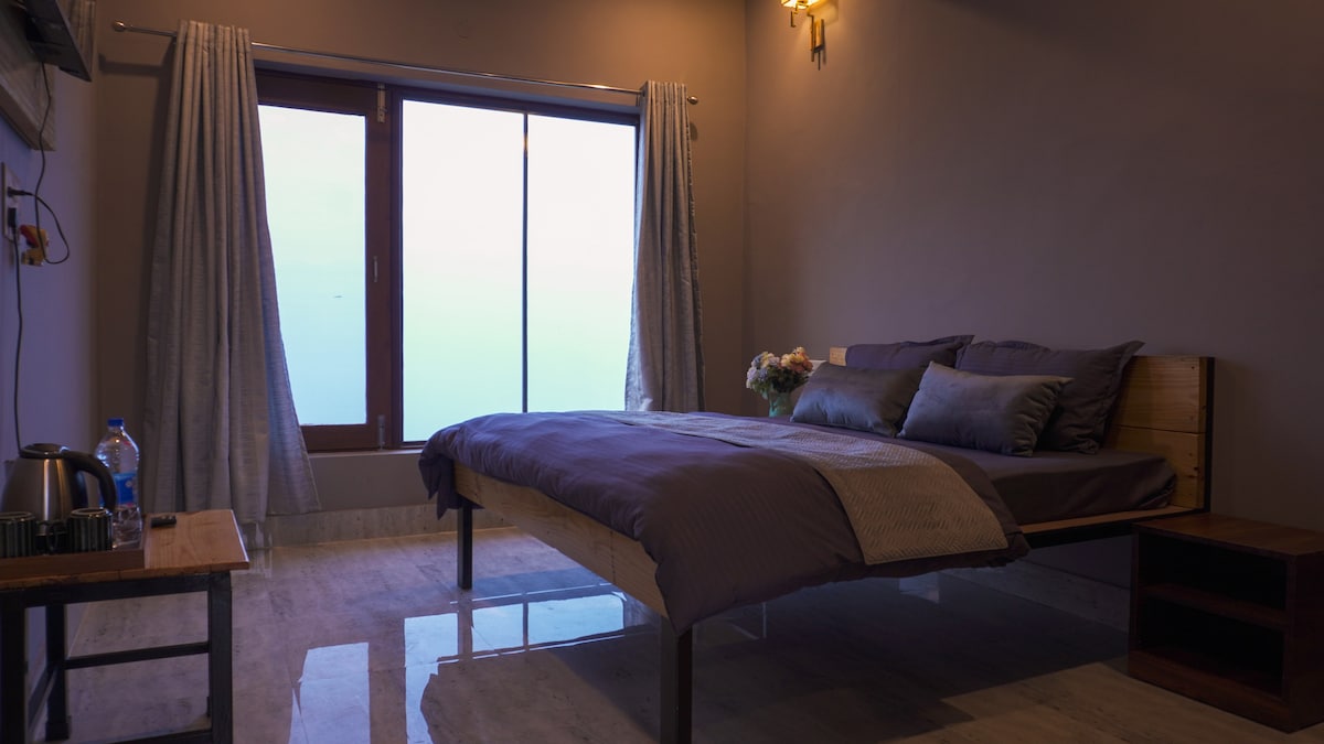 A spacious bedroom features a large bed dressed in neutral tones, with several decorative pillows. Sunlight filters through a large window with sheer curtains, providing a calming view. A small table with a kettle is positioned nearby, enhancing the room's functionality.