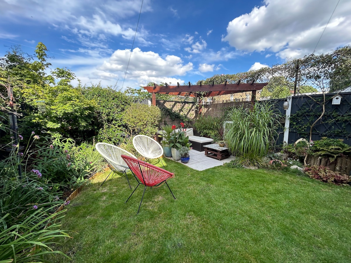 A serene garden features a well-maintained lawn with various plants and flowers. Two stylish chairs, one red and one white, are positioned on a paved area. A wooden pergola overhead is adorned with greenery, providing a cozy nook for relaxation.