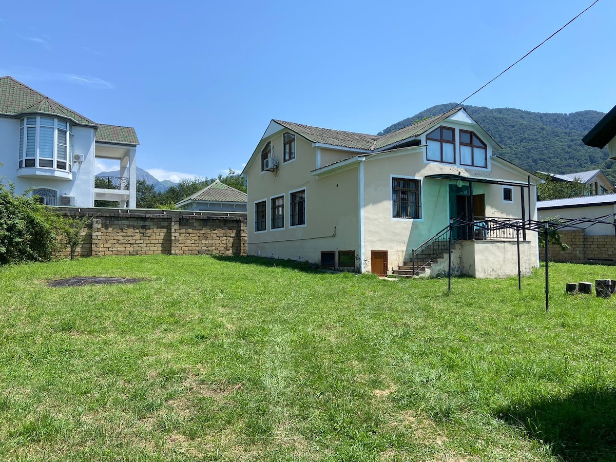 A large house is set against a backdrop of rolling hills and clear blue skies. The exterior features a mix of light-colored walls and large windows. A spacious green yard surrounds the home, providing ample outdoor space.