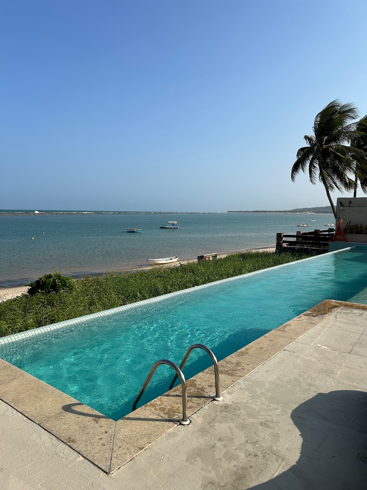 An infinity pool stretches along the edge of a serene coastal view, featuring clear turquoise water. Lush greenery and gentle waves can be seen beyond the pool, with boats floating in the distance. Palm trees sway gently under a bright blue sky.