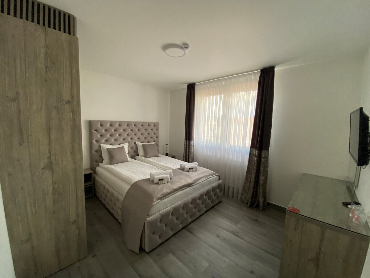 A well-appointed bedroom features a tufted headboard above a spacious bed, complemented by neutral bedding. Natural light filters through sheer curtains, illuminating the room. A wooden dresser and a wall-mounted television are positioned against adjacent walls, enhancing the functionality of the space.