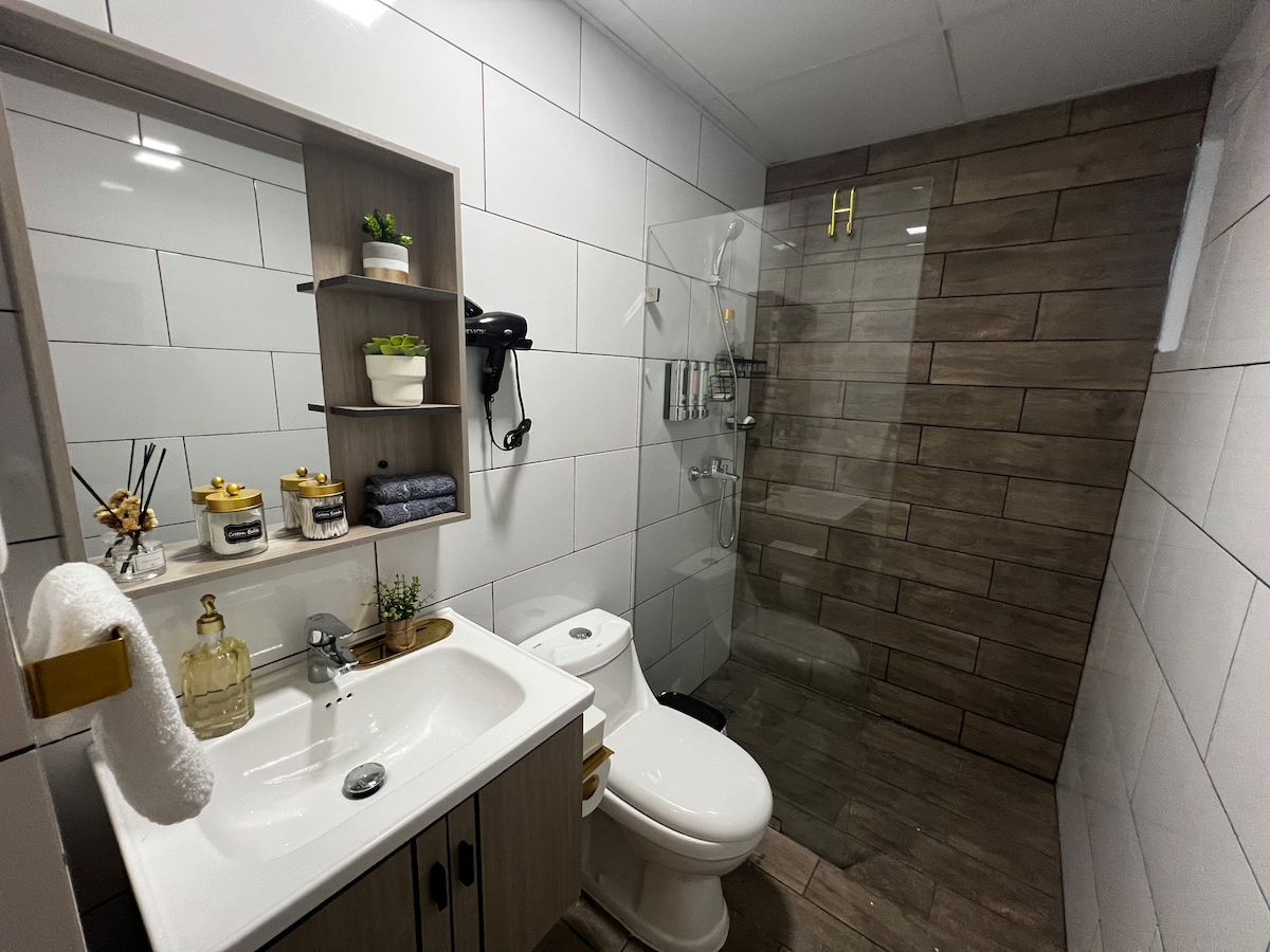 A modern bathroom features a glass shower with sleek fixtures and wood-inspired tiles. A white sink and toilet are positioned adjacent, with a wooden shelf displaying decorative items and toiletries. Natural light enhances the clean, minimalist design.