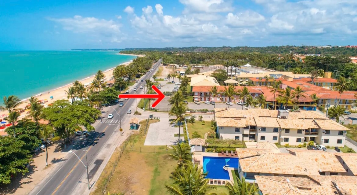 An aerial view captures the proximity of the property to Taperapuan Beach, with the coastline visible on the left and a road lined with palm trees on the right. The sparkling blue pool is positioned within the condominium grounds alongside green spaces.