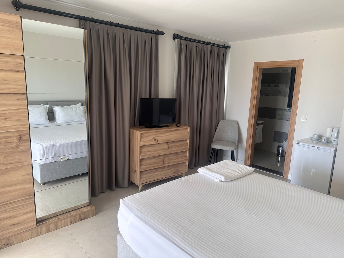 A modern hotel room features a spacious bed with crisp linens. A dresser and a small seating area are present, alongside a television mounted on the wall. Neutral tones dominate the decor, while the large curtains frame a welcoming atmosphere.