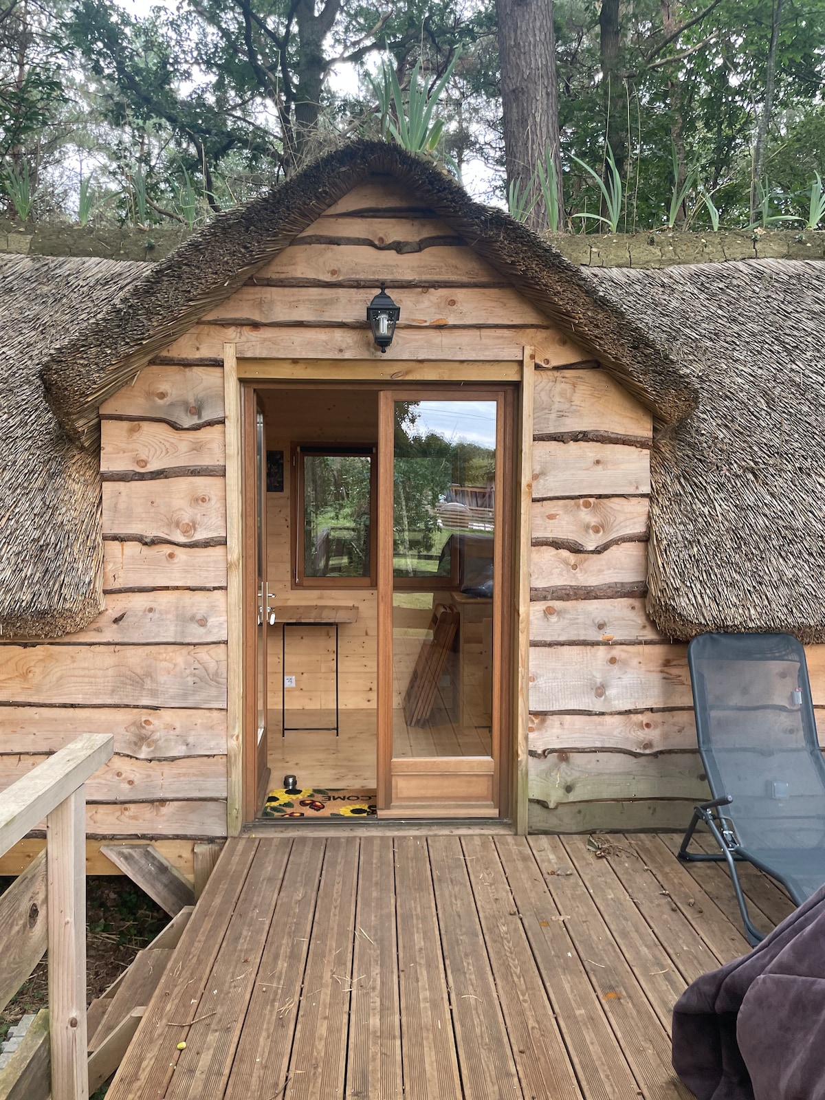 The entrance showcases a rustic wooden door framed by natural straw thatching. The welcoming space features a wooden deck with a sturdy railing, offering a glimpse of the lush greenery beyond. A cozy seating option is positioned nearby.