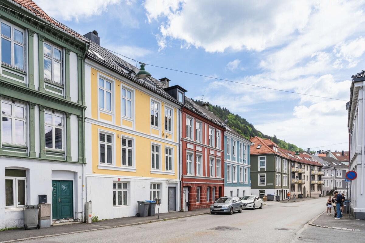 A charming street scene features a variety of buildings in different colors and styles. The facades exhibit a mix of pastel hues, including green, yellow, red, and blue, creating an inviting atmosphere. Lush green hills rise in the background, enhancing the neighborhood's character.