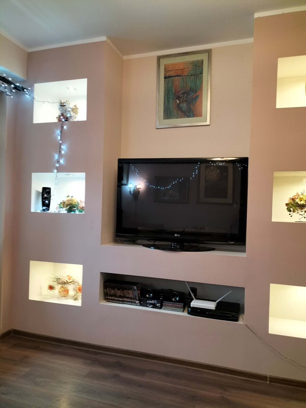 A wall-mounted smart TV is positioned within a light-toned alcove, surrounded by decorative niches containing plants and art objects. Soft lighting enhances the space, contributing to a cozy atmosphere, ideal for relaxing entertainment such as movies and music.