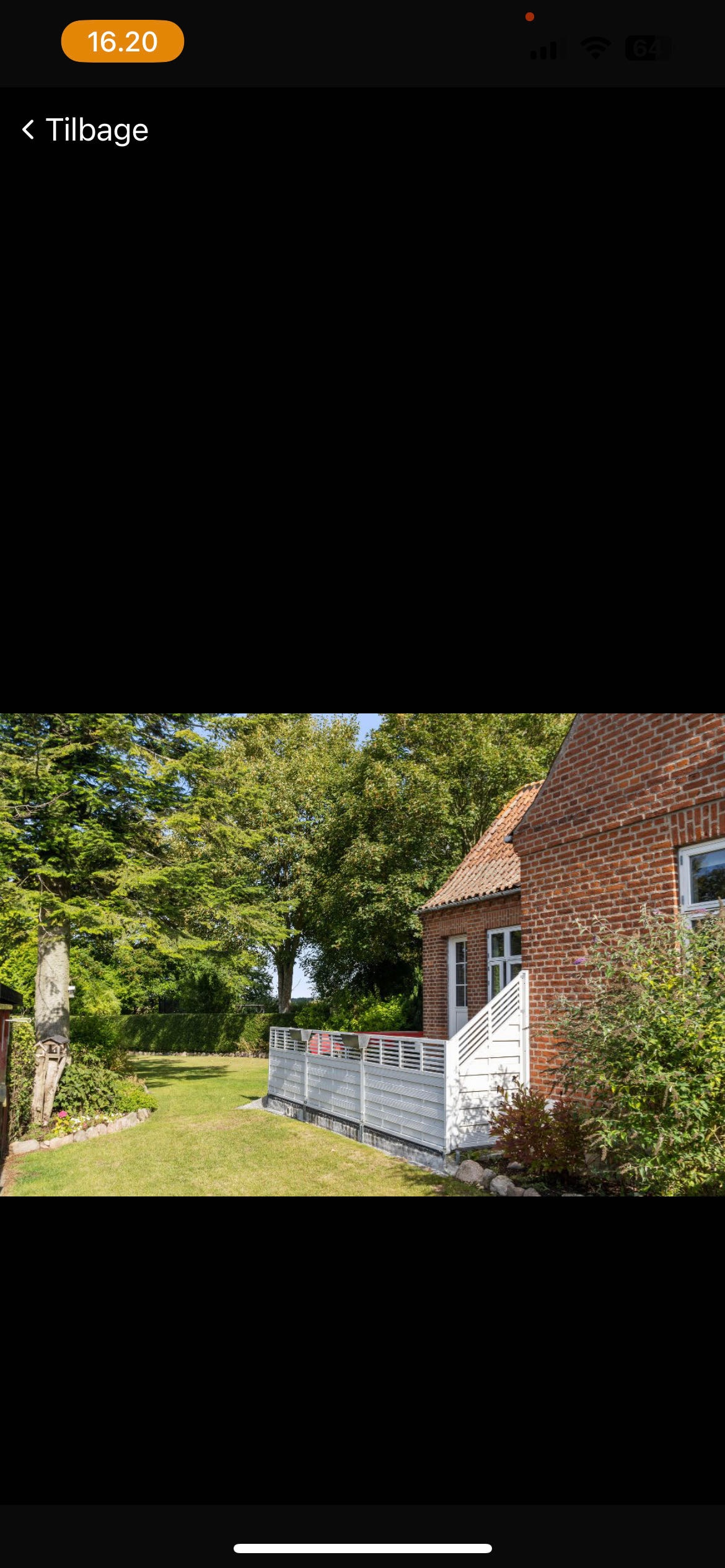 A charming exterior view highlights a brick house with a sloped roof, surrounded by lush greenery. A wooden deck is visible, leading to a well-maintained lawn that extends toward a tree-lined background, creating a serene outdoor setting.