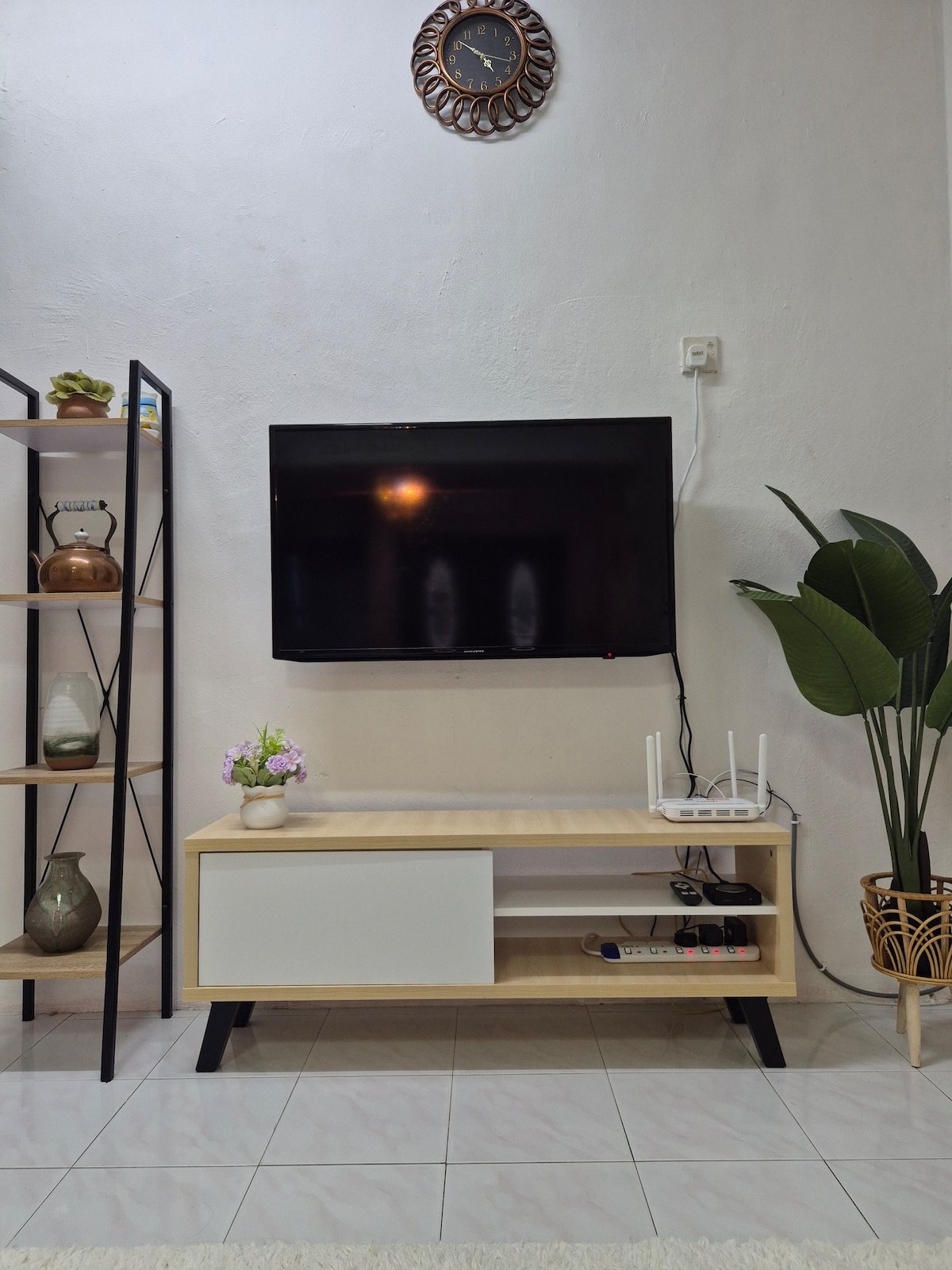 A modern living area features a sleek television mounted on a white wall. Below it, a light wood entertainment unit displays decorative items and provides storage. A decorative plant is situated next to the unit, adding a touch of greenery. A wall clock is positioned above the television.