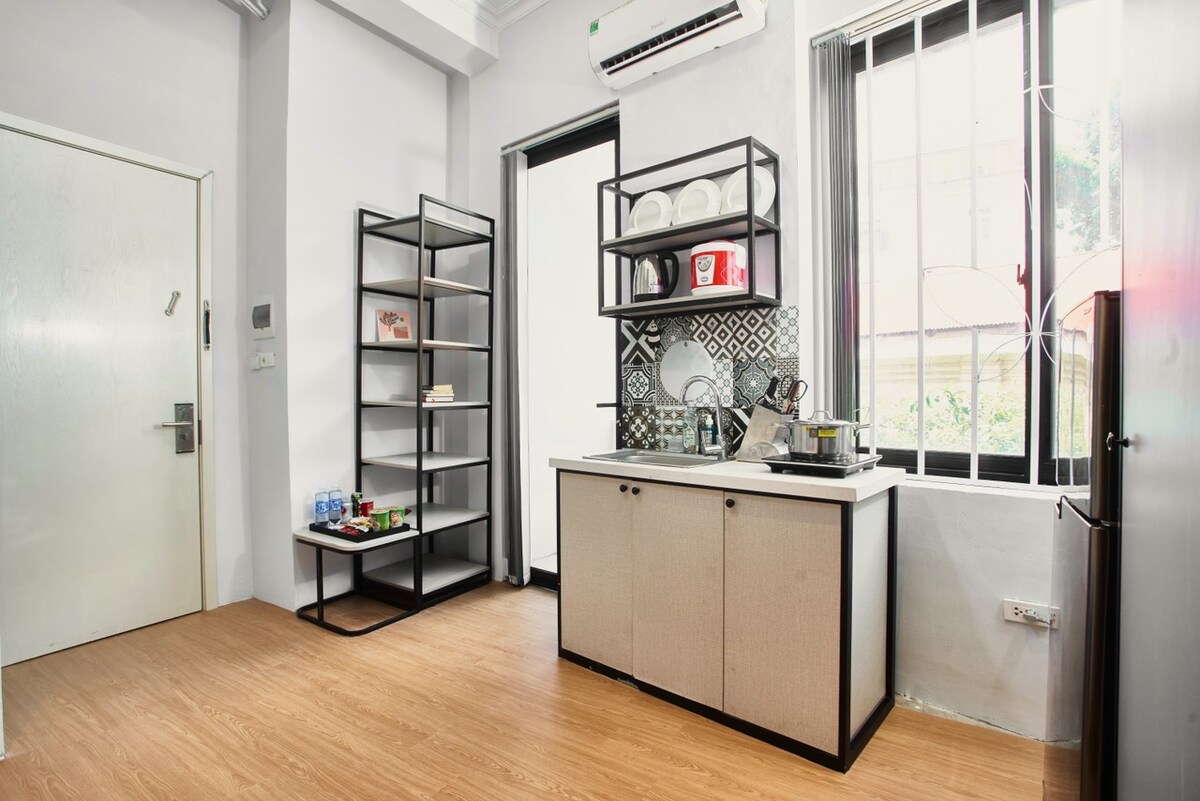 A compact kitchenette features a mounted shelf holding various dishes and utensils, with a small sink and cooking area below. A refrigerator and additional storage are present along the wall, while natural light enters through a window, enhancing the functional space.