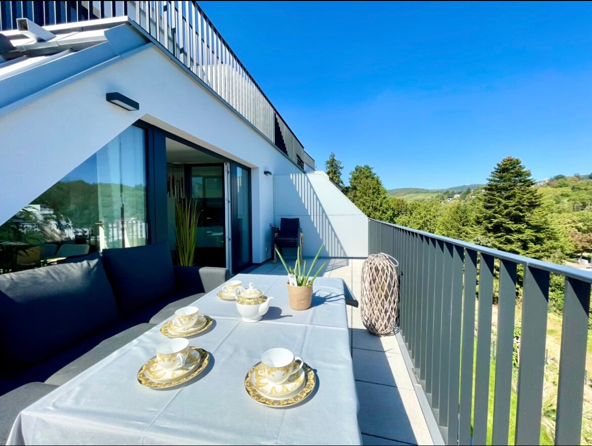 A modern balcony is presented with a dining table set with elegant dishware. A cozy seating area is visible nearby, complemented by potted plants. The large glass doors and surrounding greenery enhance the open space, with a clear blue sky visible above.