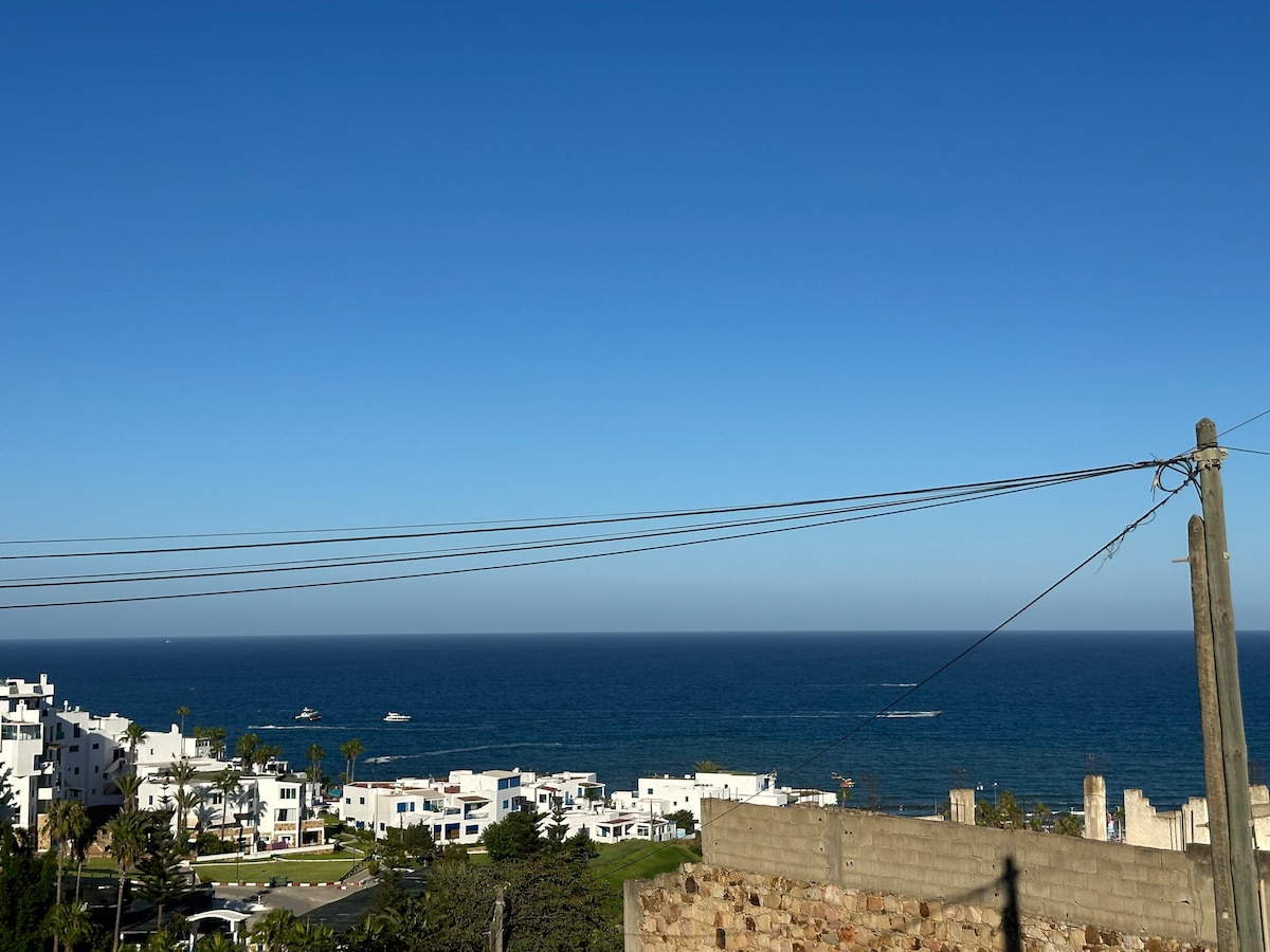 An expansive view of the ocean stretches across the horizon under a clear blue sky. The coastline features white buildings and palm trees, while boats can be seen gliding on the water. Power lines run across the foreground, framing the scene.