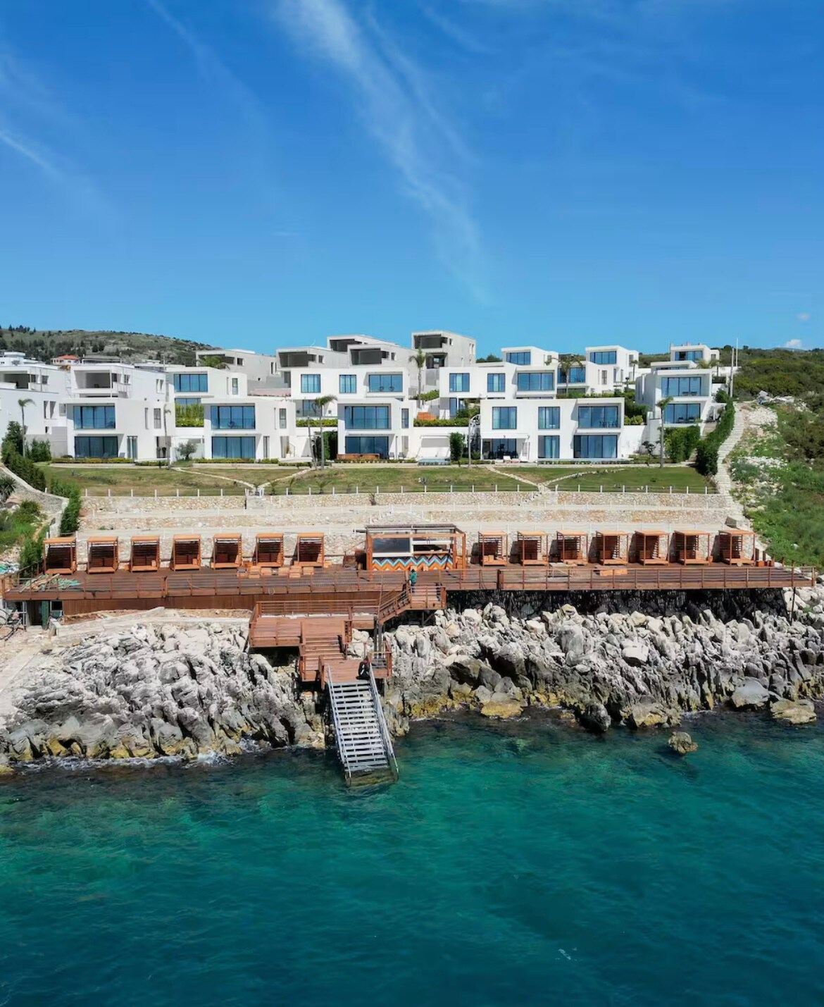 A modern villa complex is situated on rocky terrain, overlooking clear blue waters. The building features large windows and a series of wooden lounge chairs arranged on a terrace, leading down to a wooden dock extending into the water.