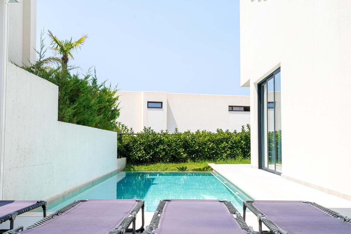 A private pool is surrounded by a sleek concrete patio, featuring several lounge chairs. Lush greenery is visible along the perimeter, offering a sense of seclusion. The clear blue sky reflects off the water's surface, enhancing the tranquil setting.