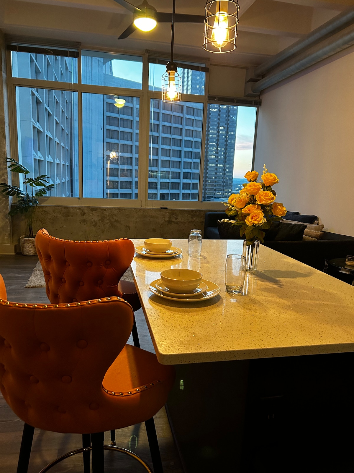 A sleek kitchen island features two orange bar stools paired with neatly arranged tableware, including plates and cups. A vase of fresh yellow roses adds a touch of color to the space, while large windows allow for ambient natural light to fill the room.